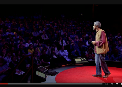 Zrzut ekranu z TED - prelegent w czerwonej koszuli, brązowej kamizelce, dżinsach i brązowych butach w czarnych butach, czarnych spodniach i grafitowym swetrze