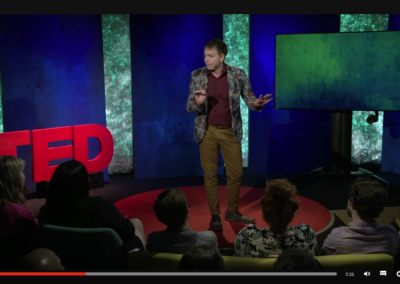 Zrzut ekranu z TED - prelegent w brązowych butach, brązowych spodniach w paski, czerwonej koszuli i marynarce w kwiatowe wzory