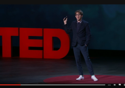 Zrzut ekranu z TED - prelegent w granatowym polo, granatowej marynarce, granatowych spodniach i białych trampkach